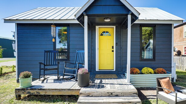 A small blue house with a yellow door, a porch with two chairs, and a planter with flowers.