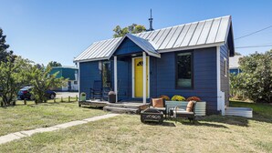 A small, single-story house with a blue exterior and a yellow door, featuring a front porch with a bench and a planter.