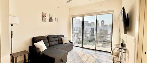 Een moderne woonkamer met een hoekbank, een staande lamp, een flatscreen televisie en een grote schuifdeur die leidt naar een balkon met uitzicht op de stad.