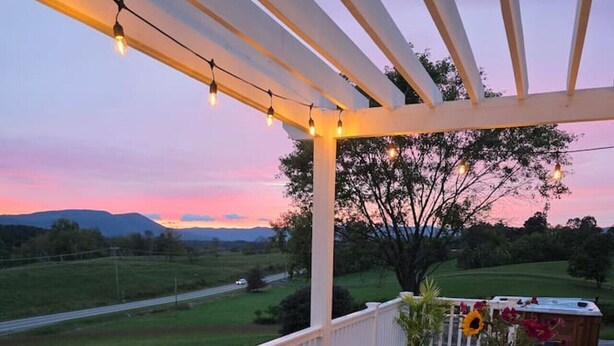 A wooden deck with a table and chairs, overlooking a grassy landscape and a pool area. - Cozy Farmhouse, Hot tub and Sauna on 8 acres! (Troutville)