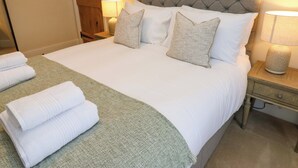 Uma cama bem arrumada com cabeceira estofada cinza, roupa de cama branca e duas luminárias de cabeceira.