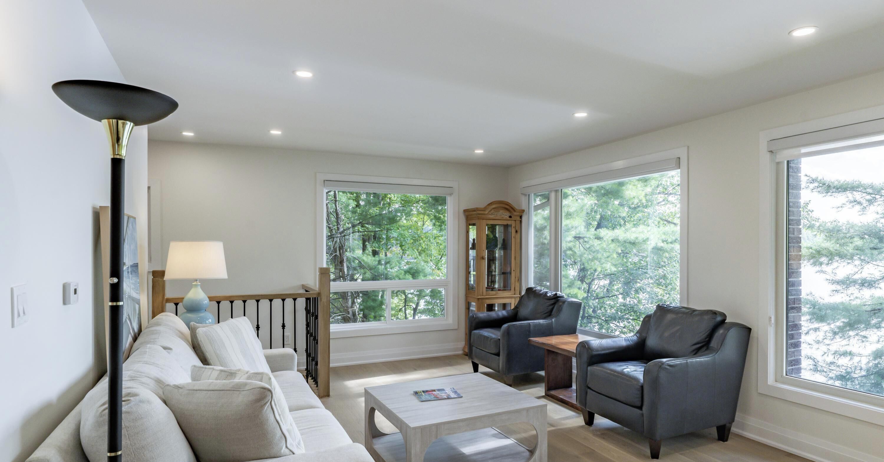 A modern living room with a sofa, two armchairs, a coffee table, and a lamp. There are large windows with a view of trees.
