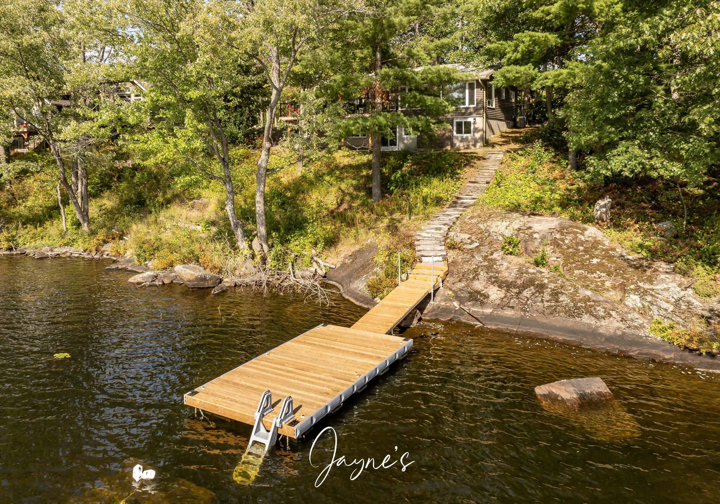 A wooden dock extending into a river surrounded by trees, with a house in the background.