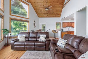 A spacious living room with a brown leather sectional sofa, a ceiling fan, and large windows offering a view of the outdoors.