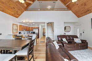 A spacious living area with a wooden ceiling, a dining table with chairs, a leather sofa with pillows, and a kitchen in the background.