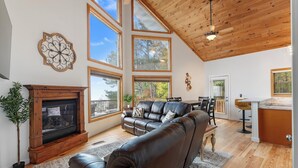 A spacious living room with a fireplace, leather sofa, dining area, and large windows offering views of trees.