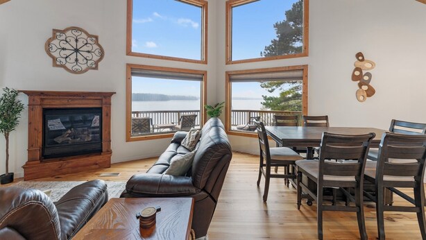 A spacious living room with a fireplace, a large sofa, a dining table with chairs, and large windows offering a view of a lake.