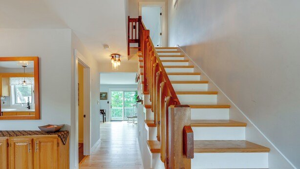 A wooden staircase with a handrail leading to an upper floor. A kitchen area with wooden cabinets and a countertop is visible on the left. - Mtn Views, On-Site Trails! Spacious Bartlett Home (Bartlett)