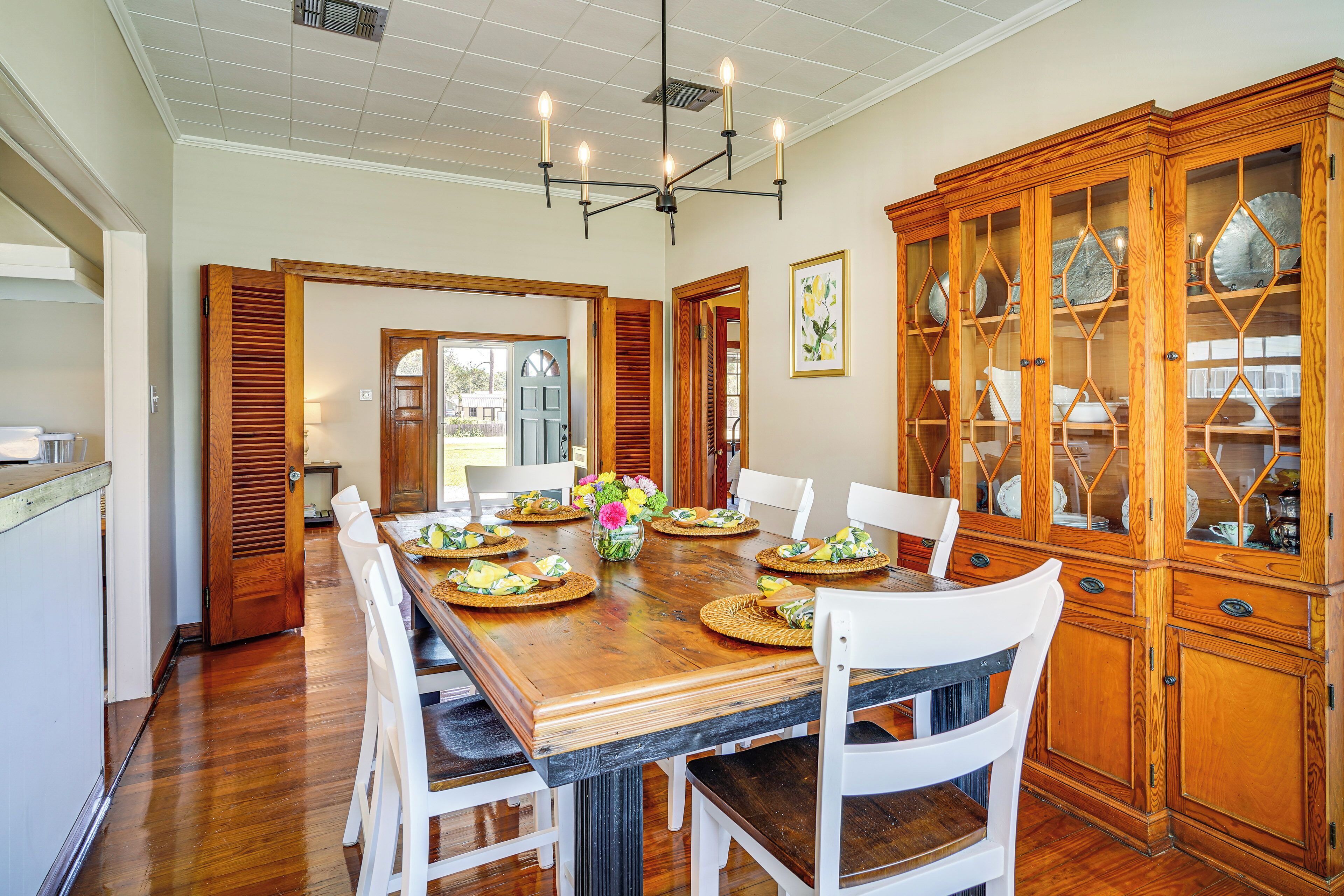 Une salle à manger avec une table en bois dressée pour six personnes, entourée de chaises blanches, et un buffet en bois avec des portes en verre sur le côté droit.