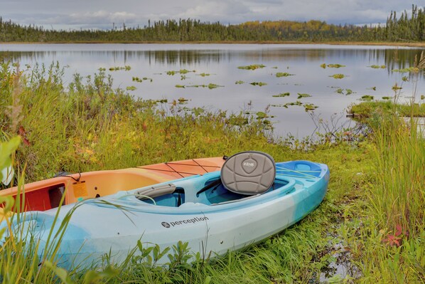 Un kayak con el nombre de la marca y una remo encima, sobre la orilla de un lago tranquilo rodeado de hierba y árboles.