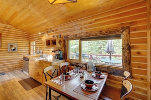 Interior de uma cabana de madeira com uma mesa de jantar posta para quatro pessoas, uma área de cozinha e uma grande janela com vista para um deck.