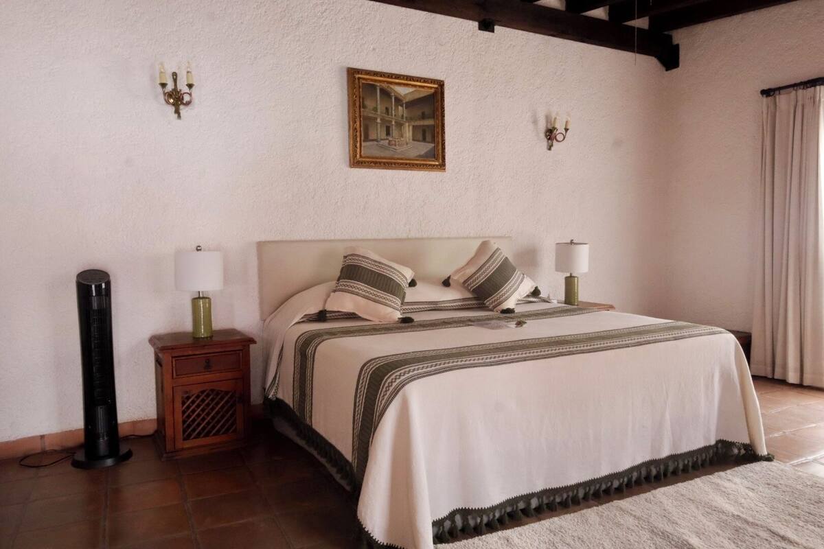Ein Schlafzimmer mit einem Bett, Nachttischlampen, einem Nachttisch, einem Bilderrahmen und einem Fenster mit Vorhängen.