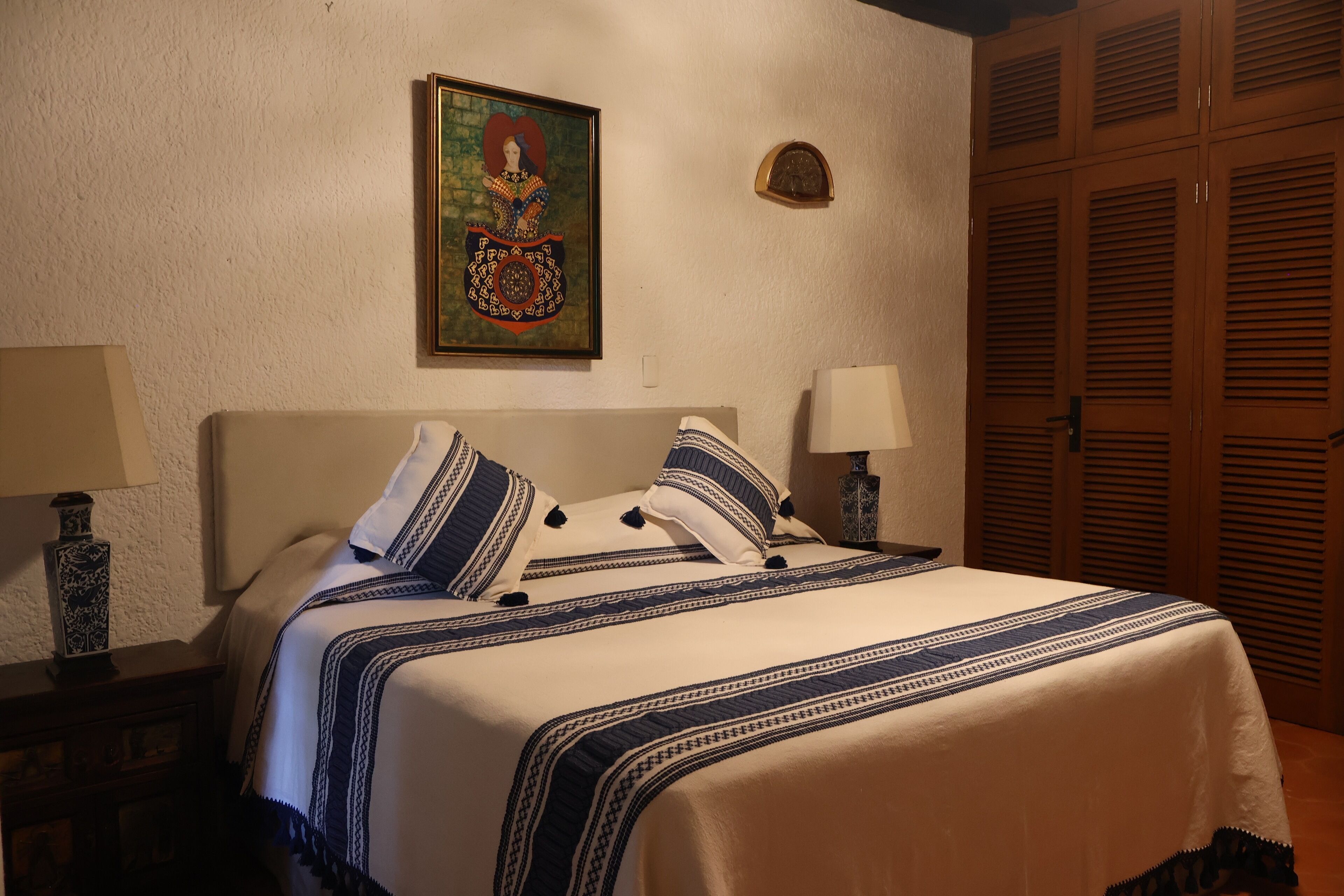 A neatly made bed with a patterned bedspread, two bedside lamps, a framed artwork on the wall, and wooden louvered closets.
