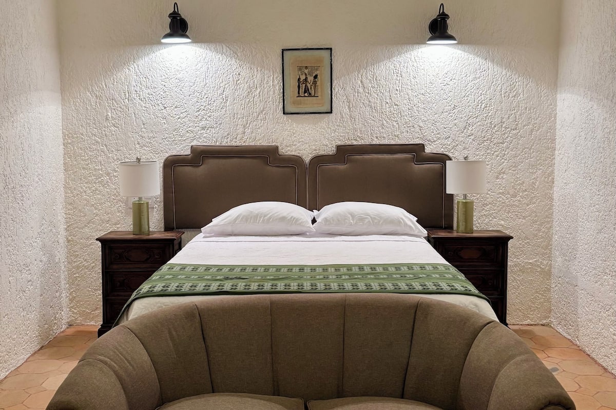 Ein Schlafzimmer mit einem Doppelbett, zwei Nachttischen mit Lampen, einem gerahmten Bild an der Wand und einem braunen Sofa im Vordergrund.