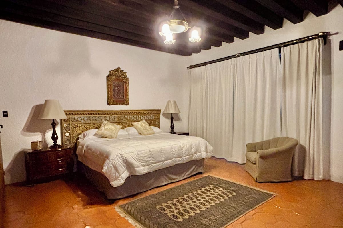 Ein Schlafzimmer mit einem Bett, zwei Nachttischen mit Lampen, einem Sessel, einem Teppich, einem Vorhang und einem an der Wand hängenden Bild.