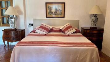 Una cama bien hecha con una colcha estampada y almohadas, flanqueada por dos mesitas de noche con lámparas y un cuadro enmarcado sobre la cama.