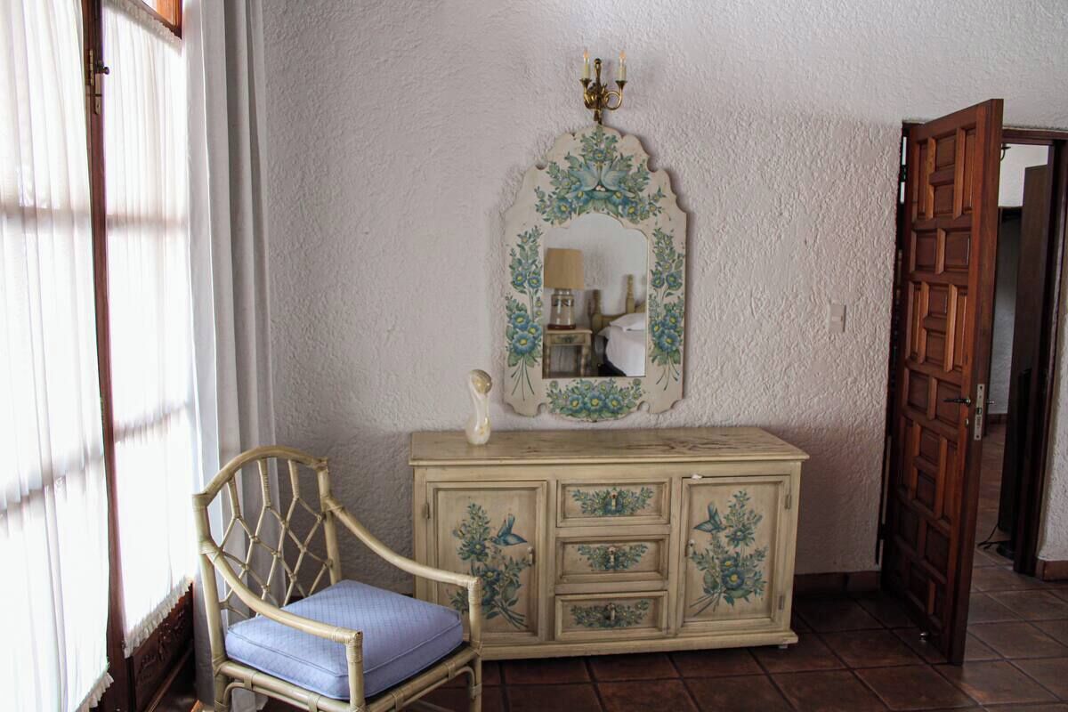 Una habitación con un armario de madera, una silla con cojín, un espejo con motivos florales y una puerta de madera.