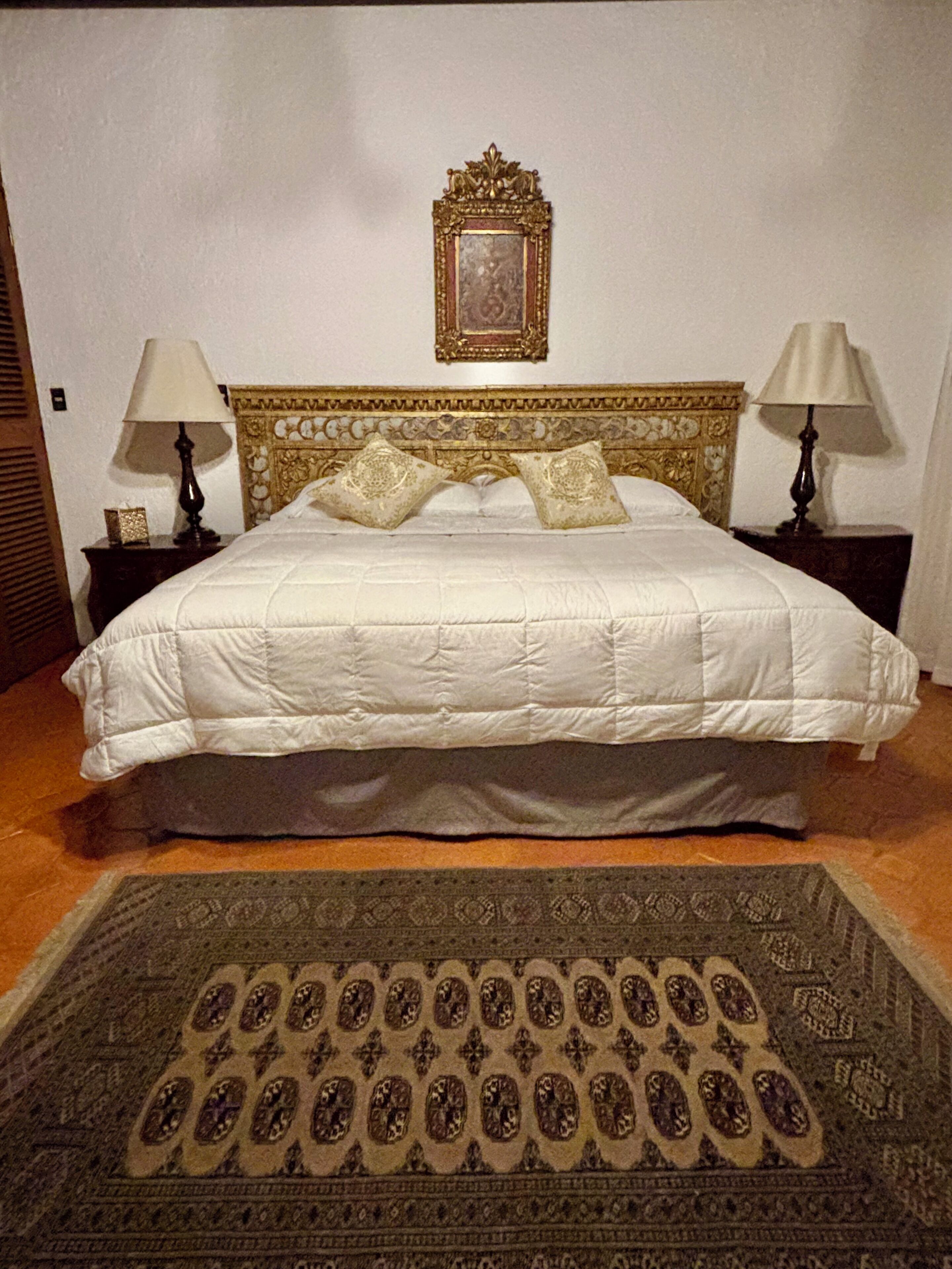 Un llit ben fet amb edredó blanc, dos coixins decoratius i un capçal amb un disseny ornamentat. Hi ha dues taules de nit amb làmpades, i una catifa amb dibuixos al terra.