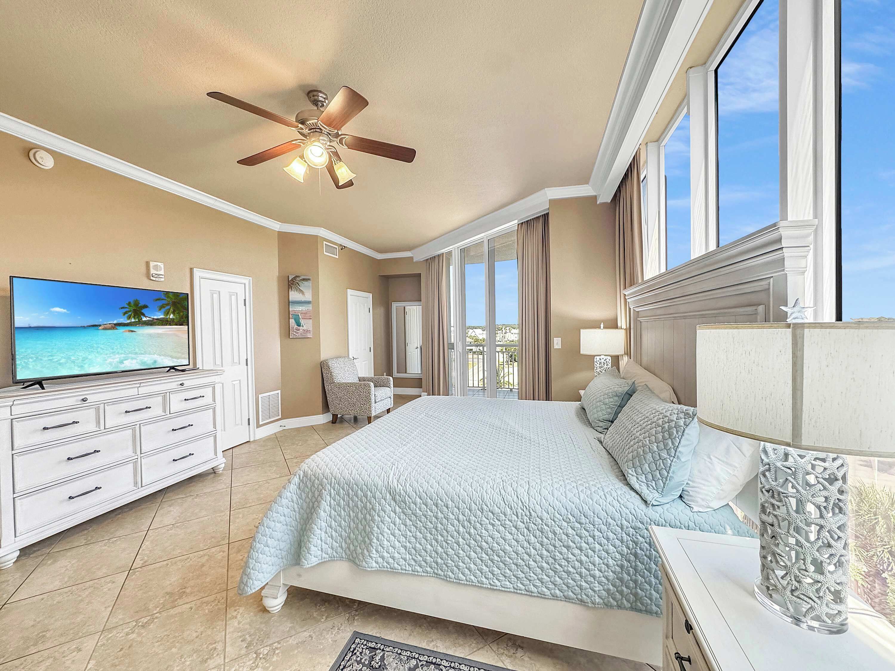 A bedroom with a bed, a ceiling fan, a TV, a chair, a desk, and a window with curtains.