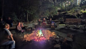A group of people sitting around a campfire in a forested area with benches and a picnic table.