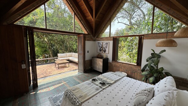 Quarto com cama grande, um sofá e vista para o exterior através de amplas janelas.