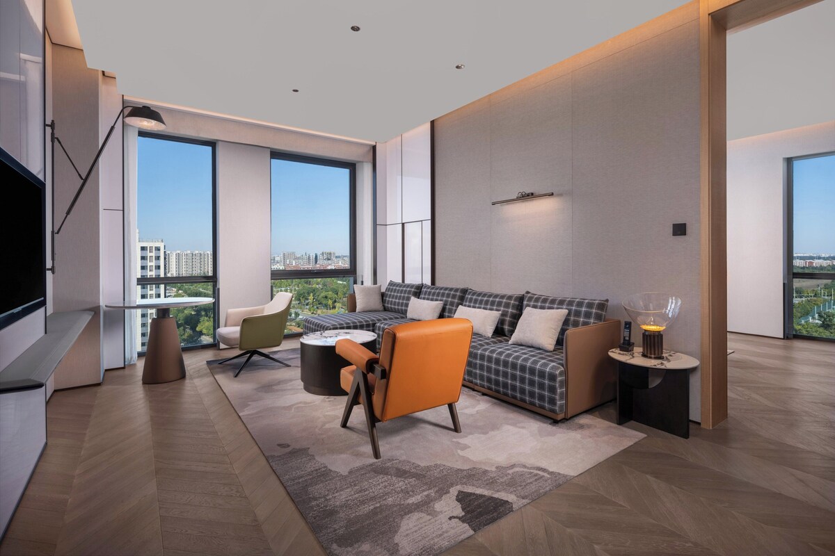 A modern living room with a large sofa, a coffee table, and a TV. There are large windows offering a city view.