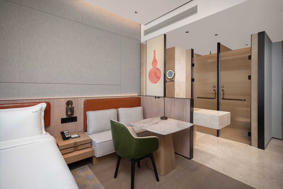 A modern hotel room with a bed, a desk, a chair, a nightstand, a mirror, and a bathroom with a sink and toilet.