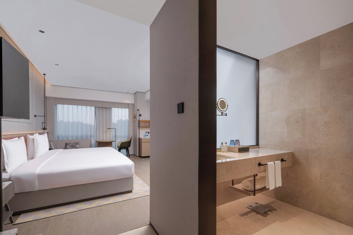 A modern hotel room with a large bed, a desk, a vanity with a mirror, and a view of the city through the window.