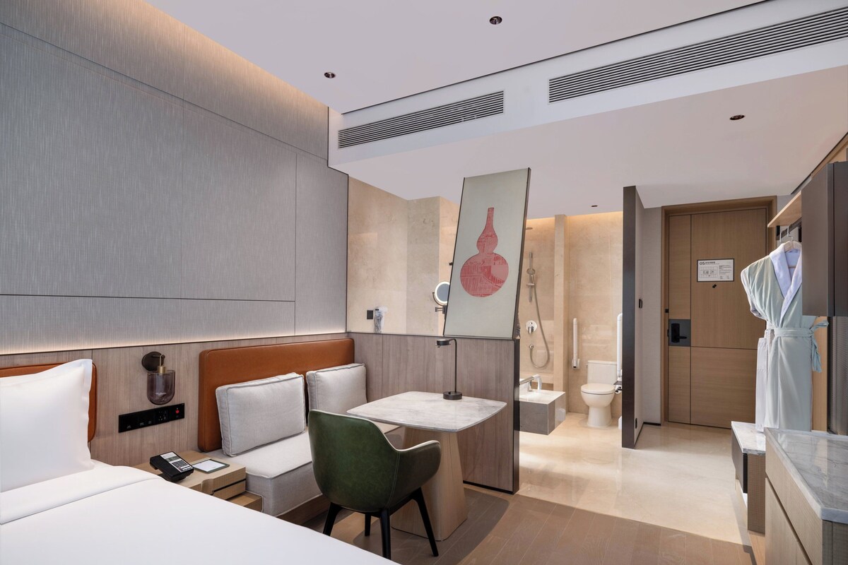 A modern hotel room with a bed, a desk, a chair, a bathroom, and a robe hanging on a rack.