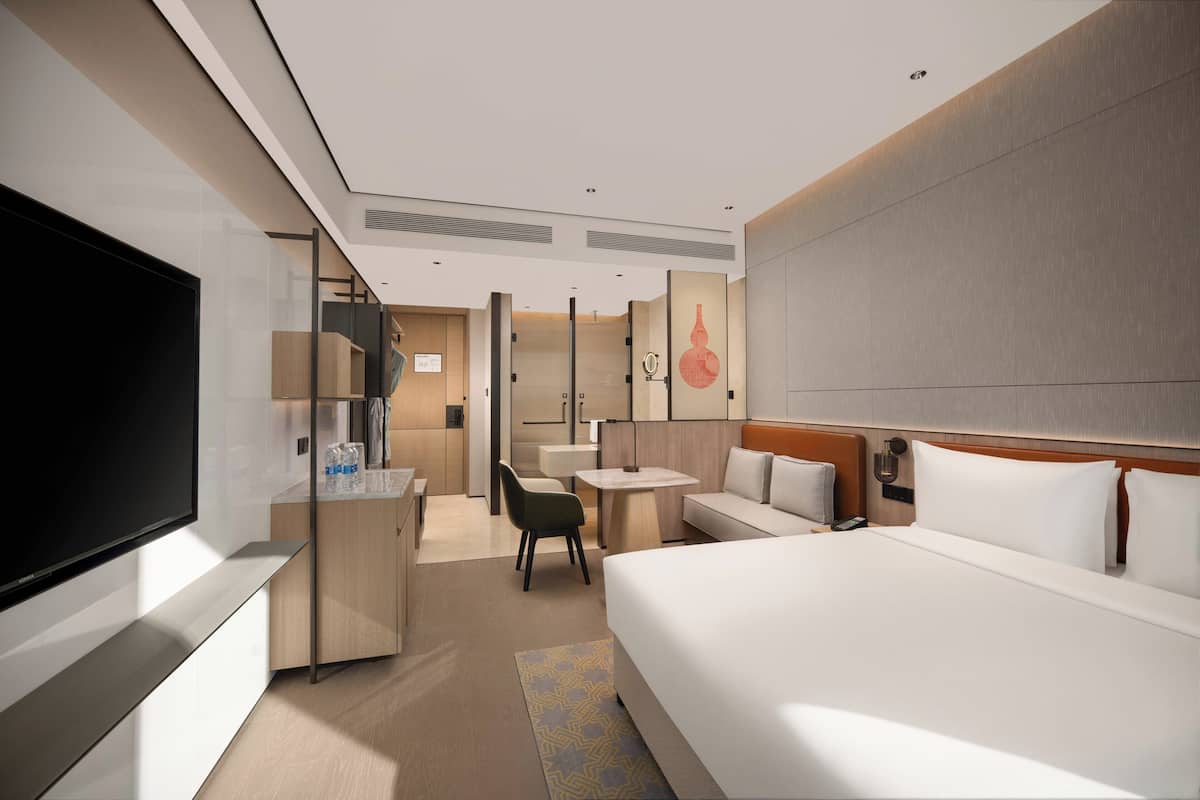 A modern hotel room with a large bed, a flat-screen TV, a desk with a chair, and a small table.