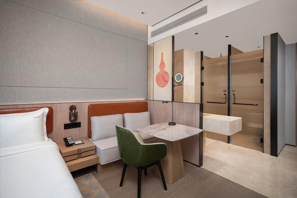 A modern hotel room with a bed, a desk, a chair, a nightstand, a mirror, and a bathroom with a sink and toilet.