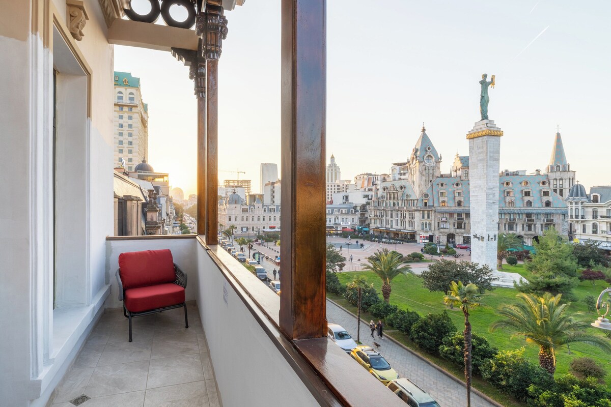 Un balcon avec une chaise rouge, donnant sur un paysage urbain composé de bâtiments historiques et d’une statue.