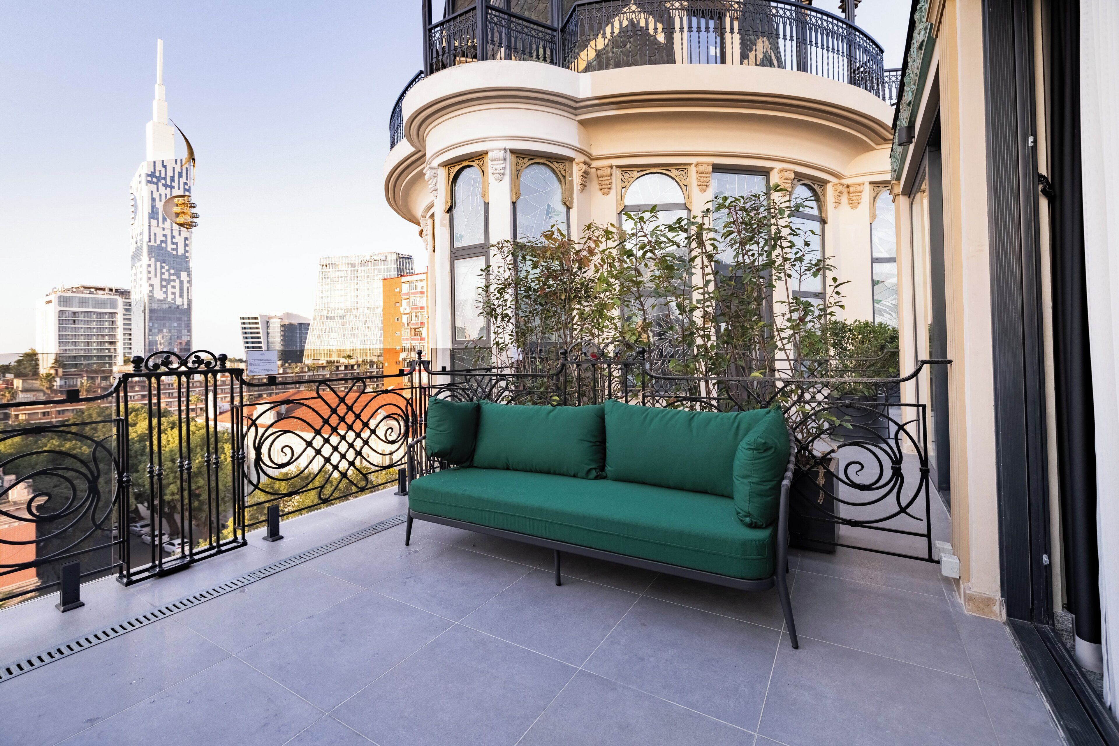 A balcony with a green sofa, ornate black railings, and a view of a cityscape including a tall building.