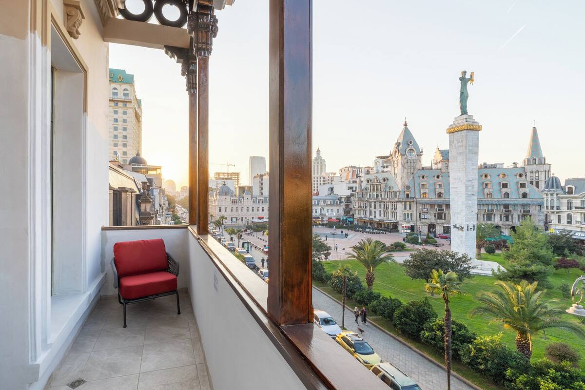 Ein Balkon mit einem roten Stuhl, mit Blick auf eine Stadtlandschaft mit historischen Gebäuden und einer Statue.