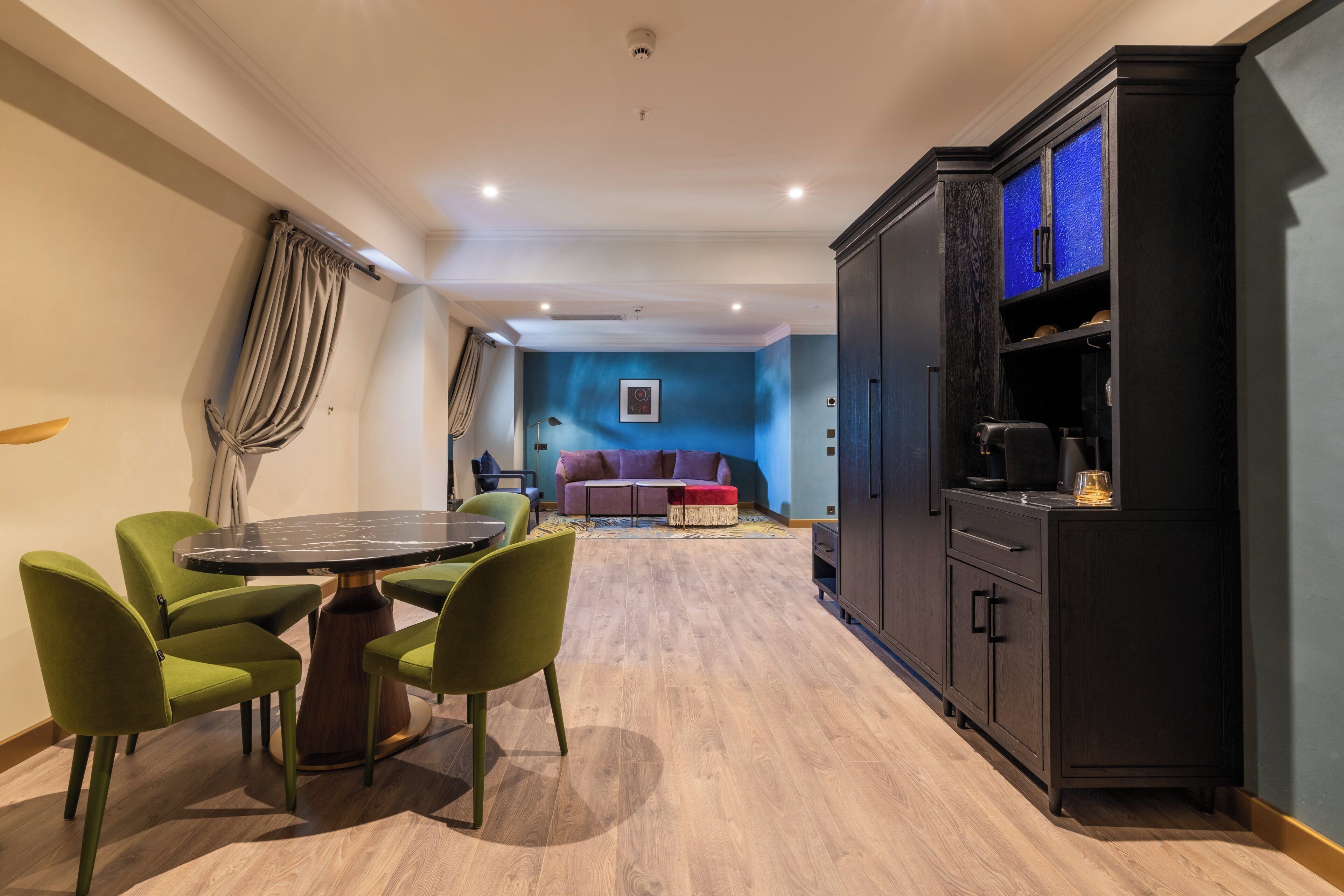 Una sala de estar moderna con una mesa redonda y sillas verdes, un mueble de madera oscura con estantes, y una pared azul decorativa.