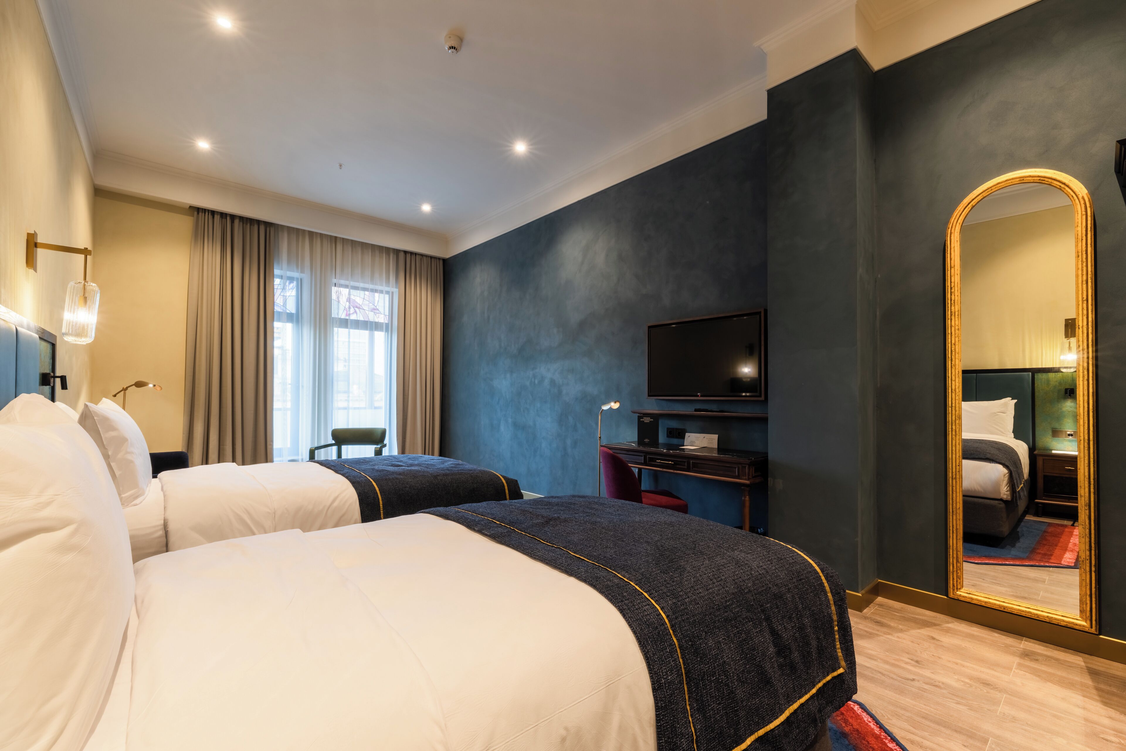 A hotel room with two beds, a TV, a desk, a mirror, and a window with curtains.