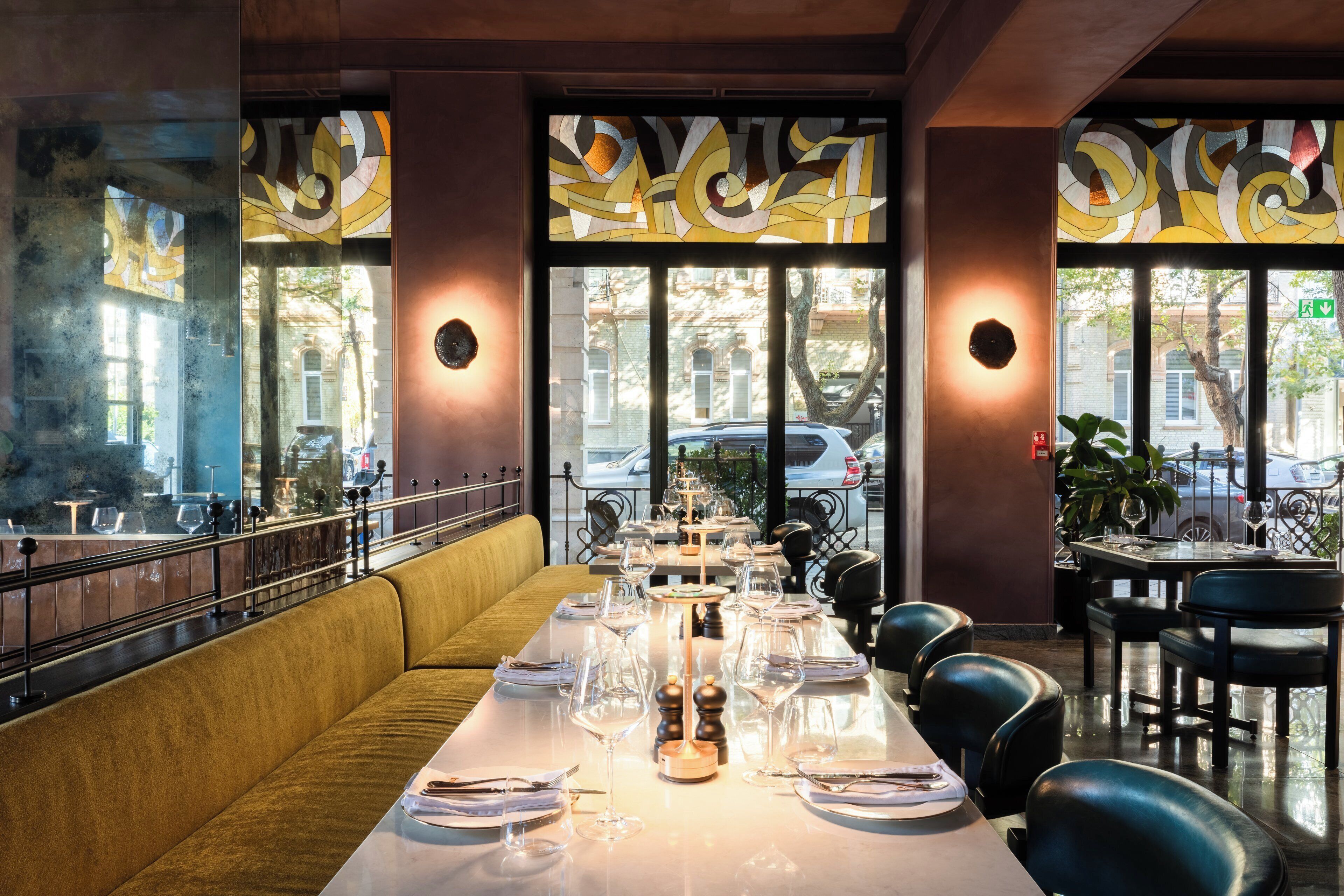 Un restaurant avec une longue table à manger dressée pour le repas, de grandes fenêtres ornées de vitraux décoratifs et une vue sur la rue.
