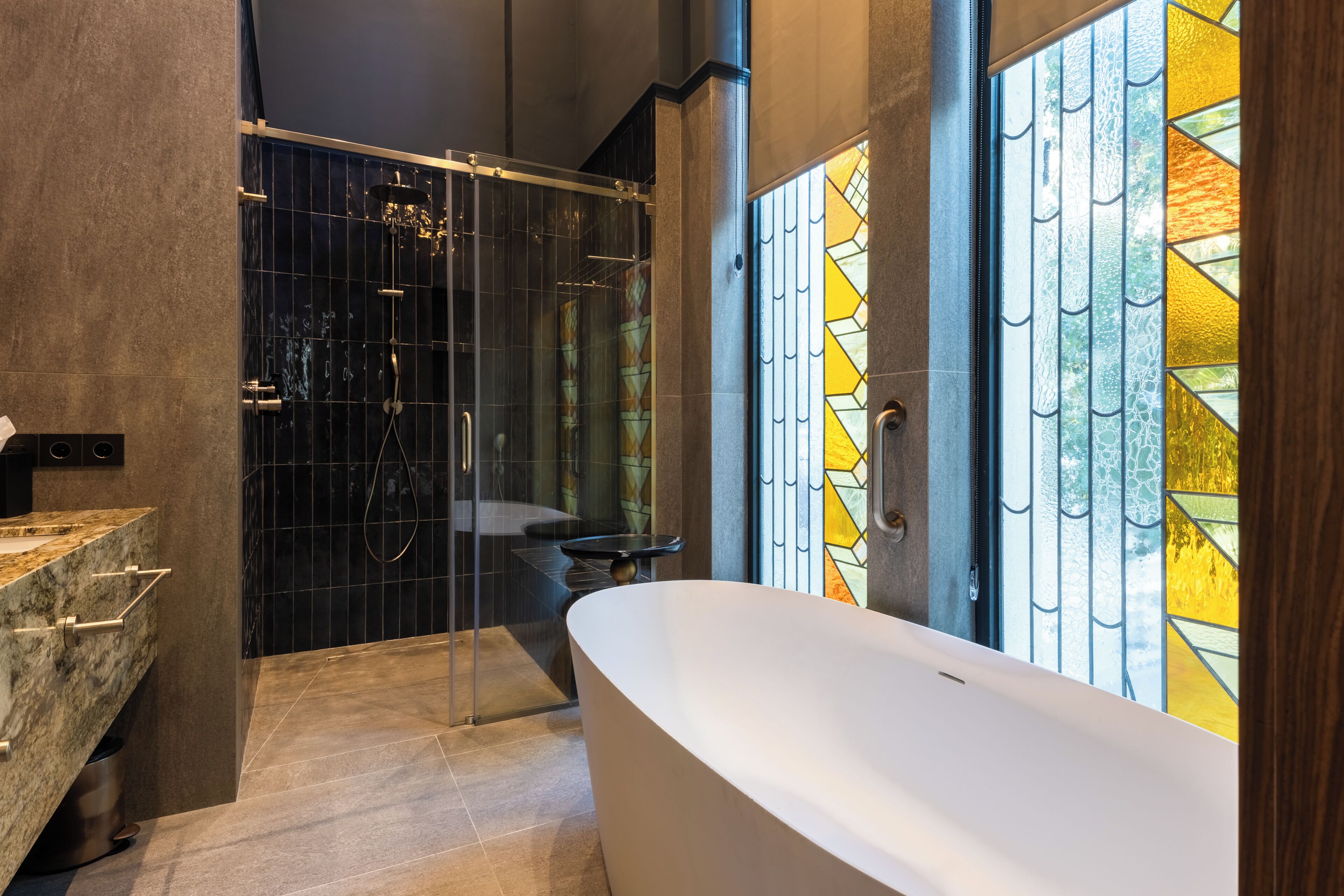 Een moderne badkamer met een vrijstaand bad, een douche met glazen wanden en een groot raam met glas-in-lood.