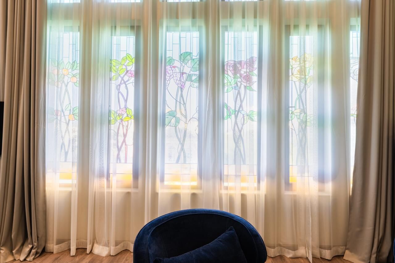 A room with a large window, sheer curtains with floral patterns, a dark blue chair, and a wooden floor.