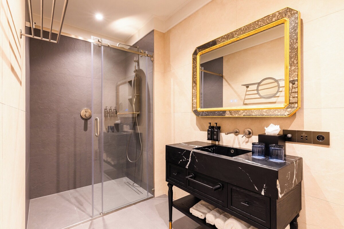 Une salle de bain moderne dotée d’une douche à parois de verre, d’un meuble-lavabo noir agrémenté de détails en marbre et d’un grand miroir avec un cadre doré.