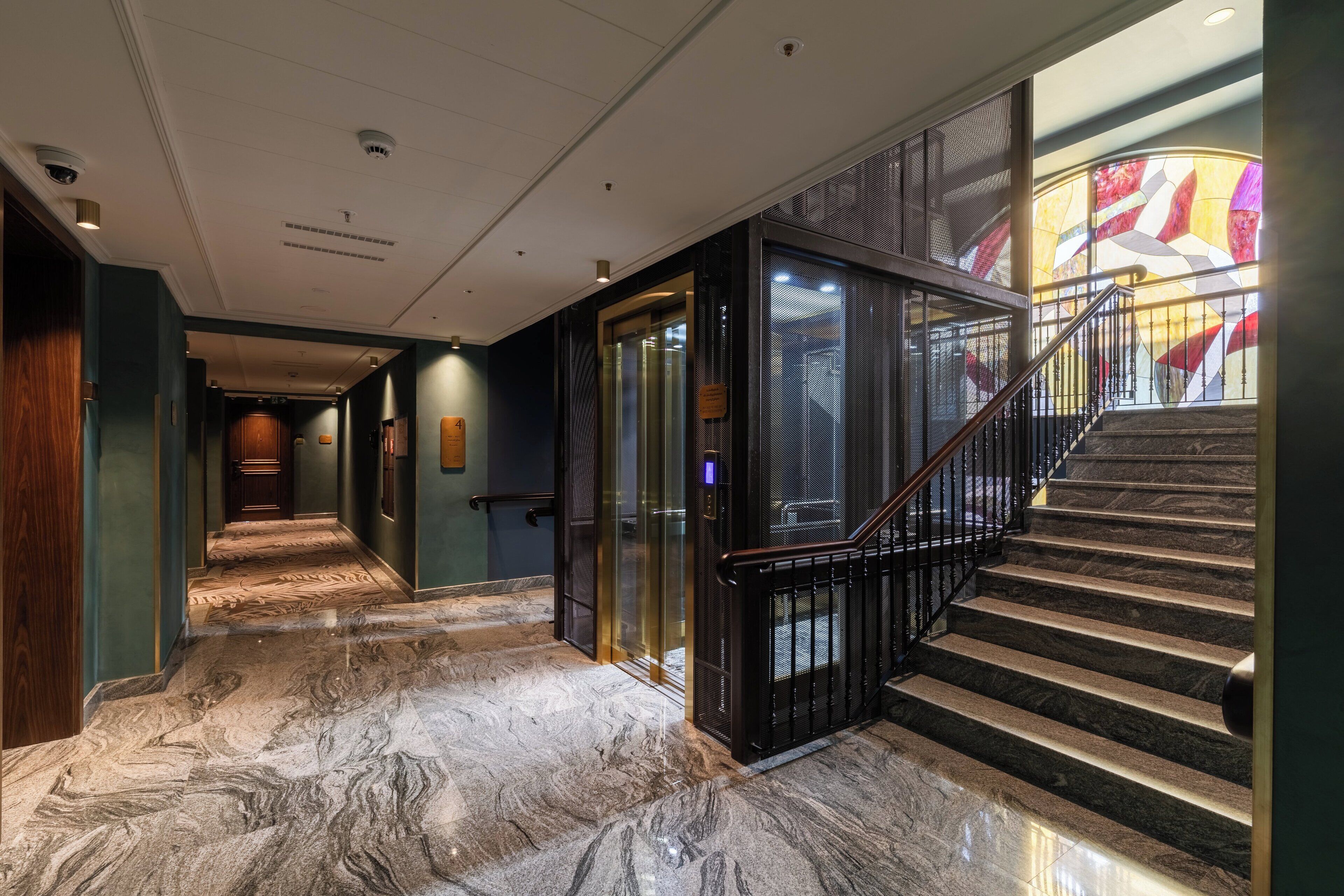A marble-floored hallway with a glass elevator, a staircase with black railings, and a large stained glass window.