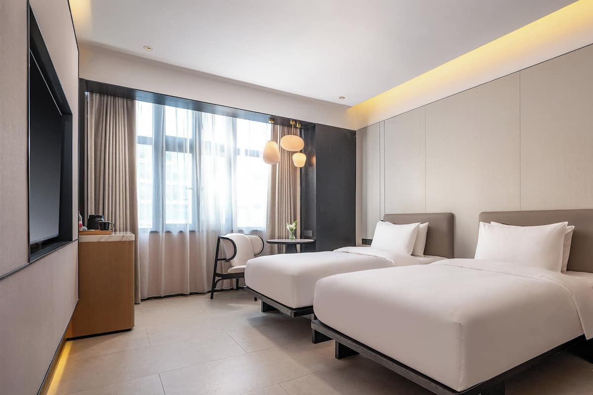 Ein modernes Hotelzimmer mit zwei Betten, einem Schreibtisch und einem großen Fenster mit Vorhängen.