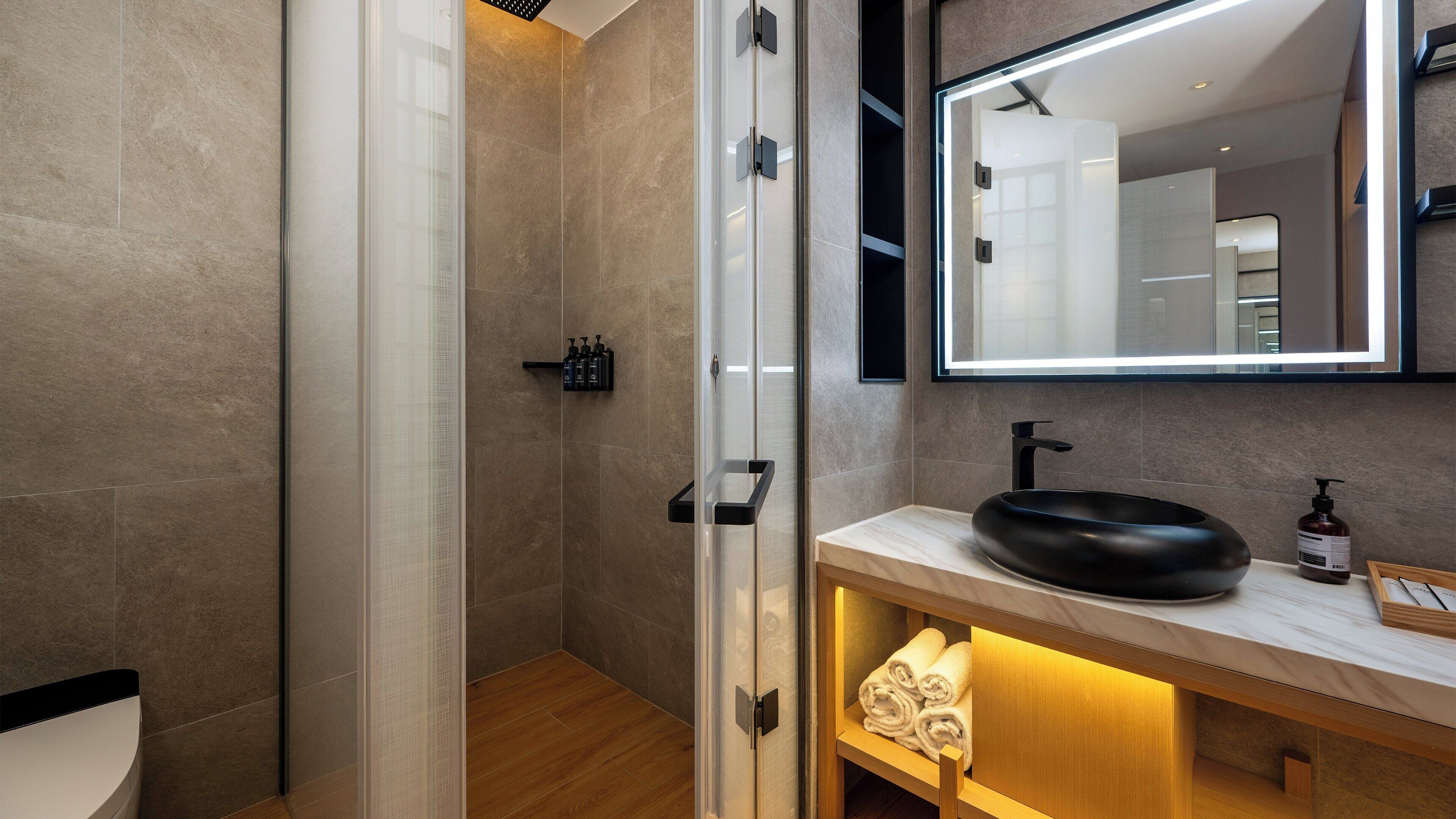 Un baño moderno con ducha envuelta en vidrio, un lavabo de diseño negro y un mueble de tocador de madera con espejo.
