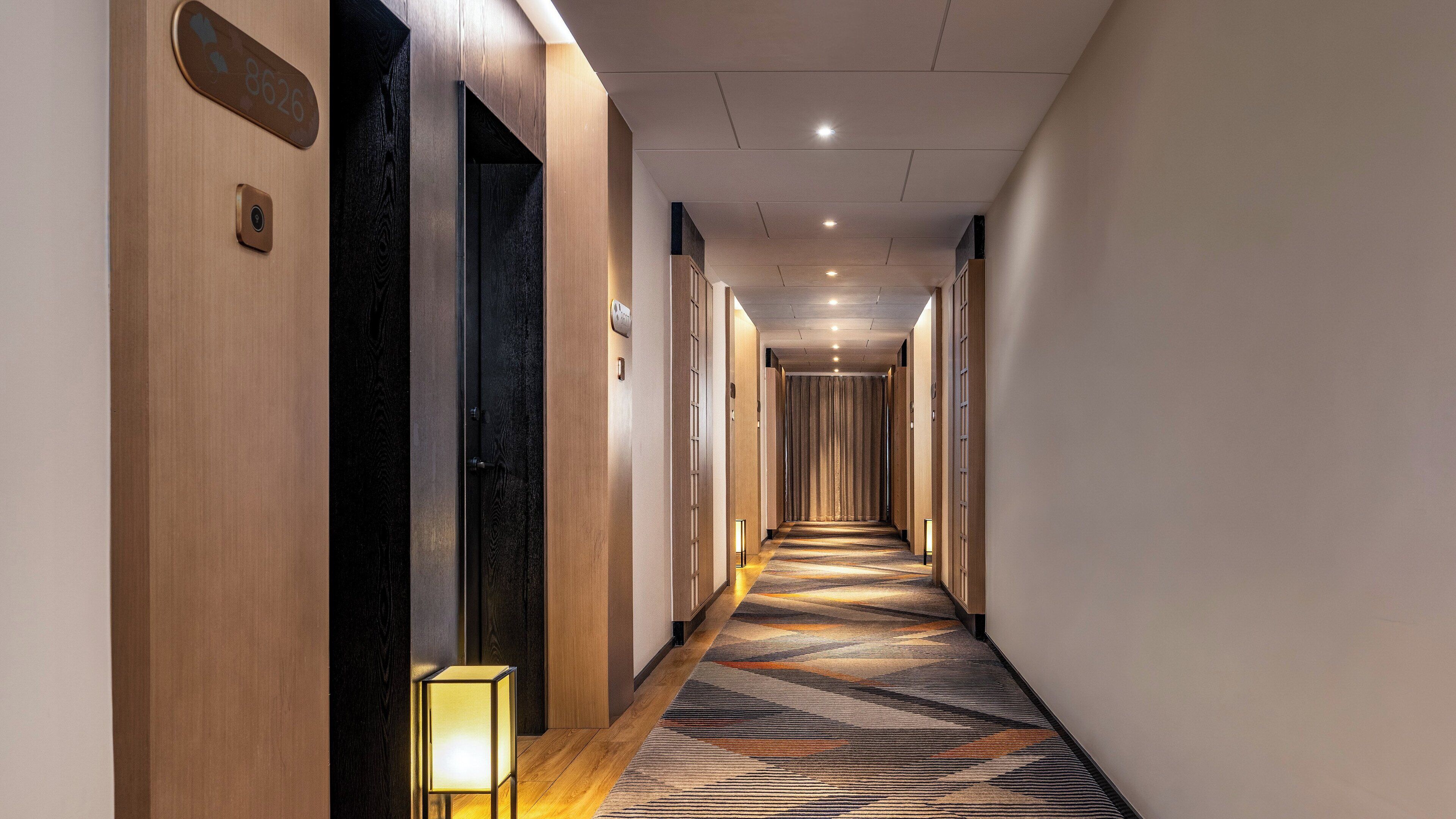 A long, narrow hotel corridor with wooden walls, a patterned carpet, and a small table lamp.