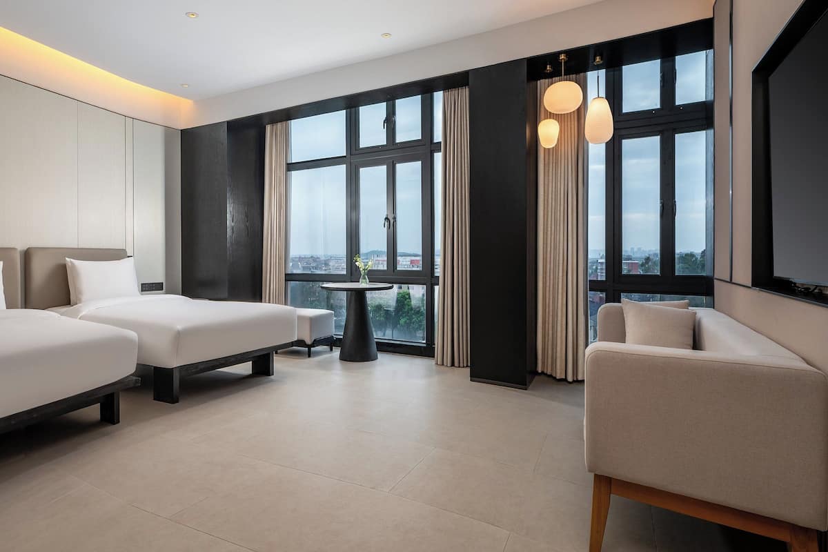 Ein Hotelzimmer mit zwei Betten, einem Schlafsofa, einem kleinen Tisch und großen Fenstern mit Vorhängen.