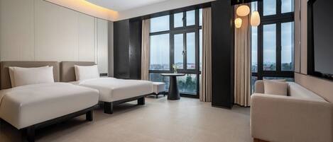 A hotel room with two beds, a sofa, a small table, and large windows with curtains.
