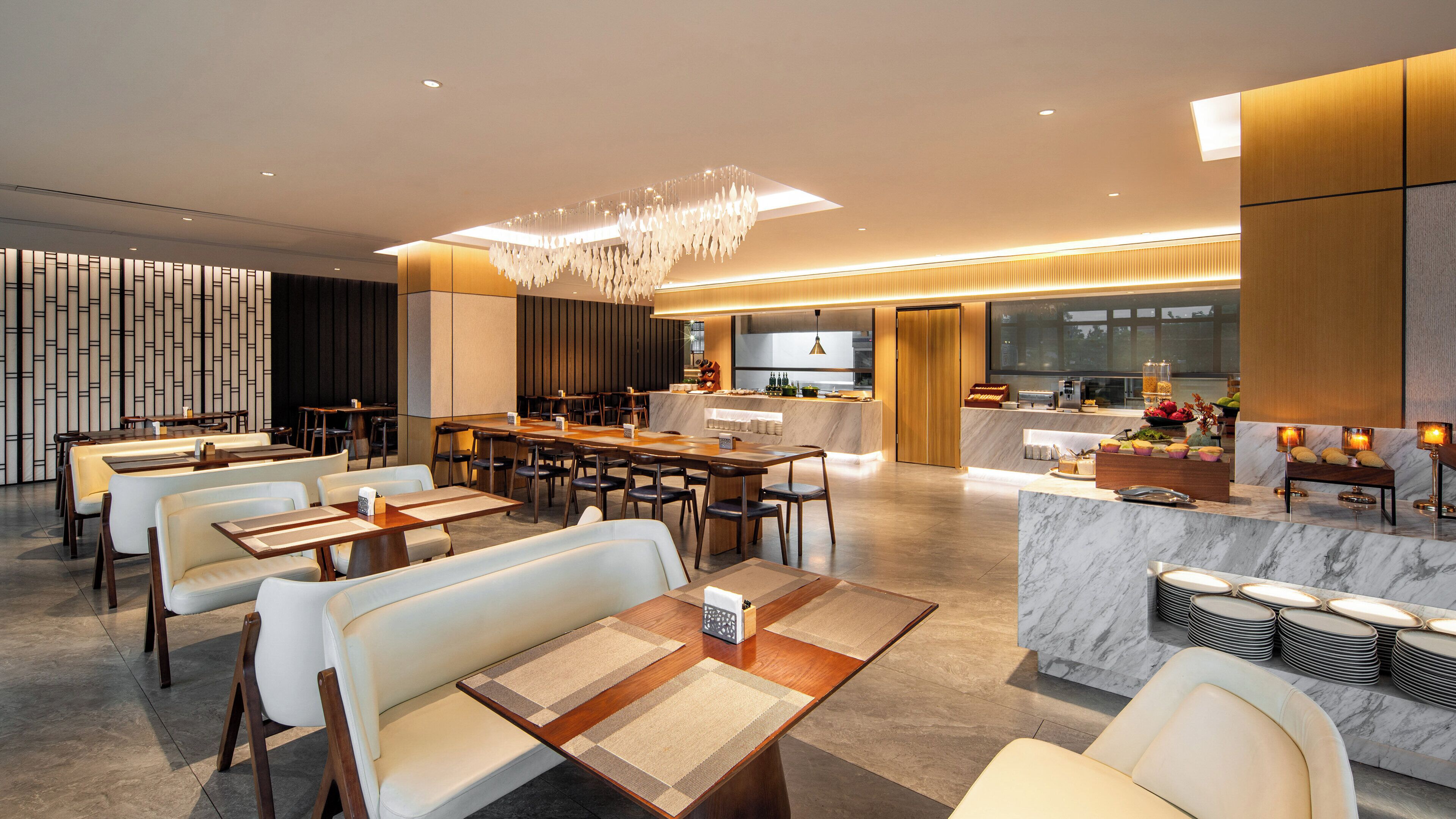 A modern hotel lobby with a dining area, a bar, and a kitchen in the background.