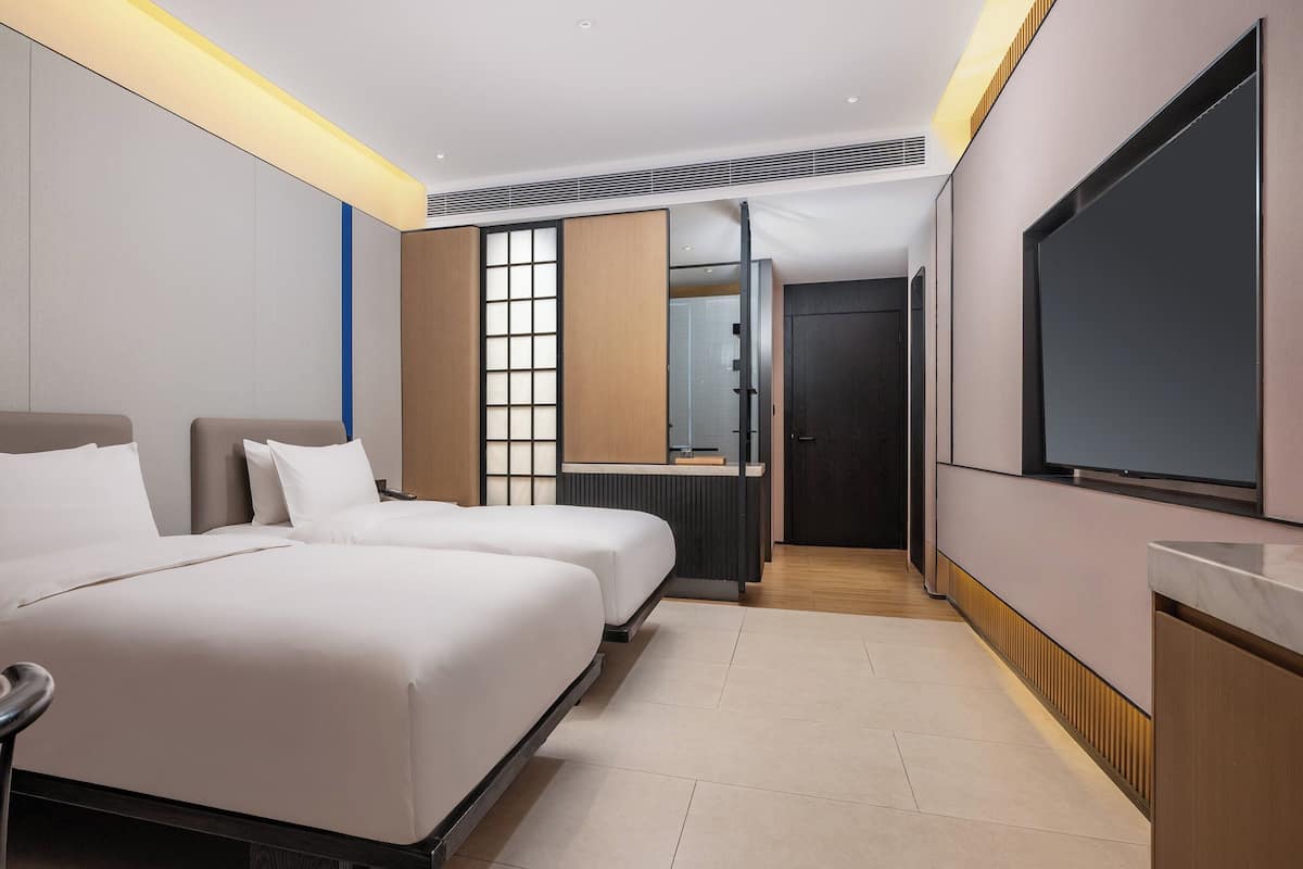 Ein Hotelzimmer mit zwei Betten, einem Fernseher, einem Schreibtisch und einem durch eine Schiebetür einsehbaren Badezimmer.