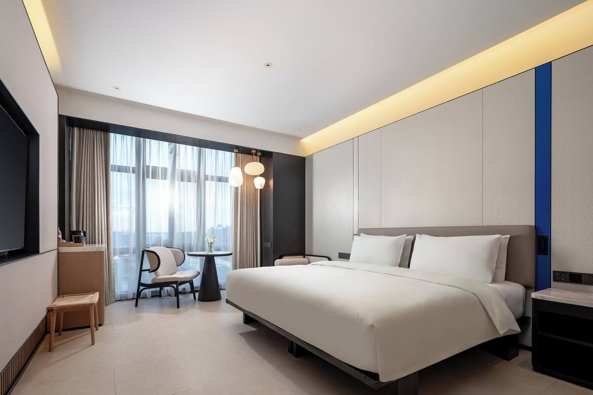 Ein modernes Hotelzimmer mit einem großen Bett, einem Schreibtisch mit Stuhl, einem kleinen Tisch ebenfalls mit Stuhl und einem großen Fenster mit Vorhängen.
