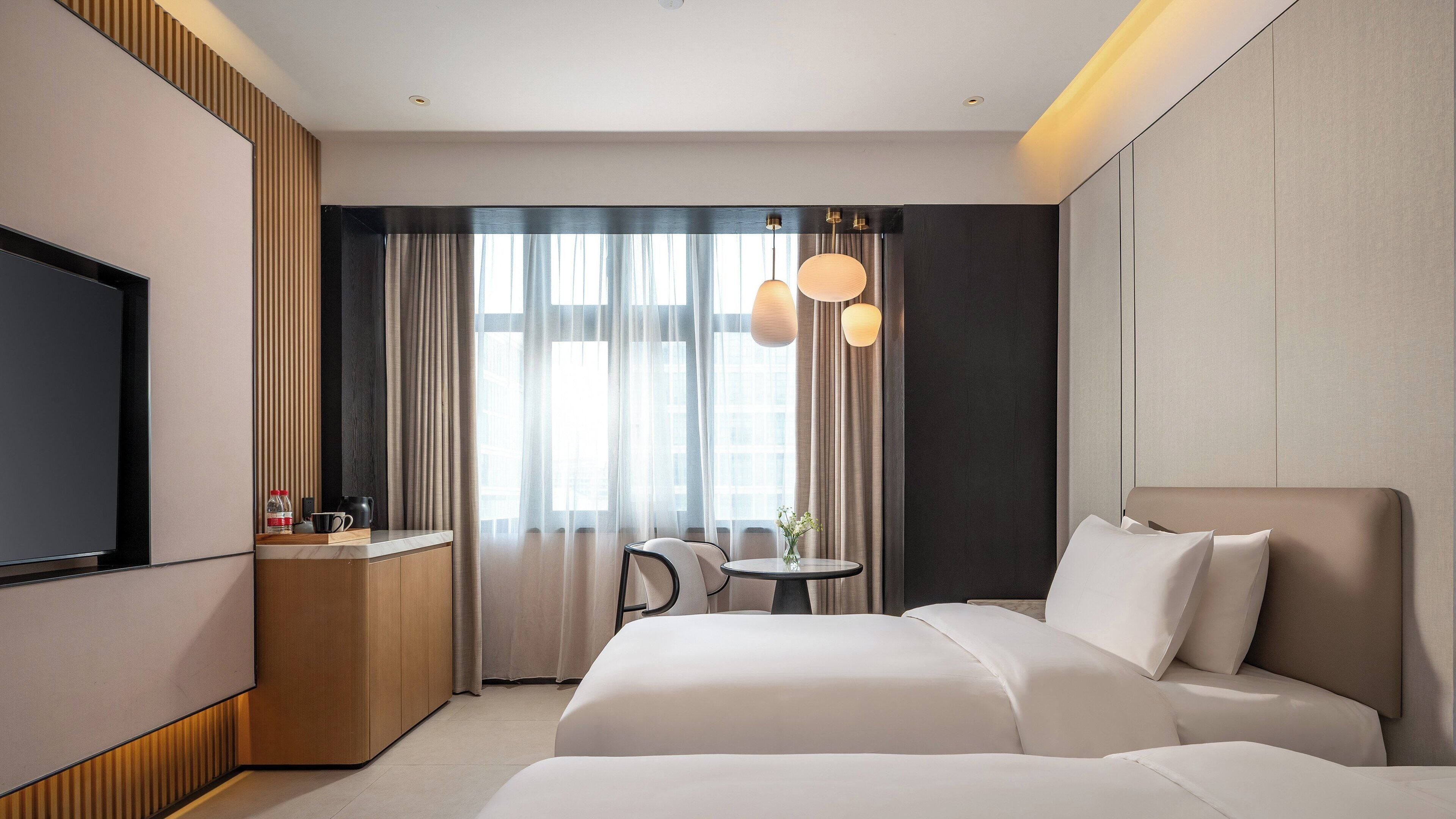 Una moderna camera d'albergo con un letto grande, una scrivania con televisore a schermo piatto, un tavolino rotondo con un vaso di fiori e vista sulla città attraverso ampie finestre con tende leggere.